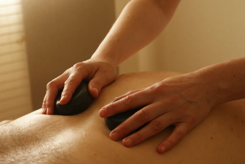 Massage aux pierres chaudes : deux mains posent des galets noirs sur le dos d'une personne allongée.